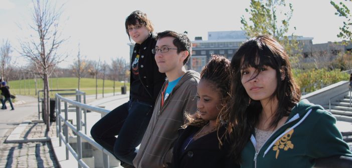 Four young people are leaning against a railing at a bus station.